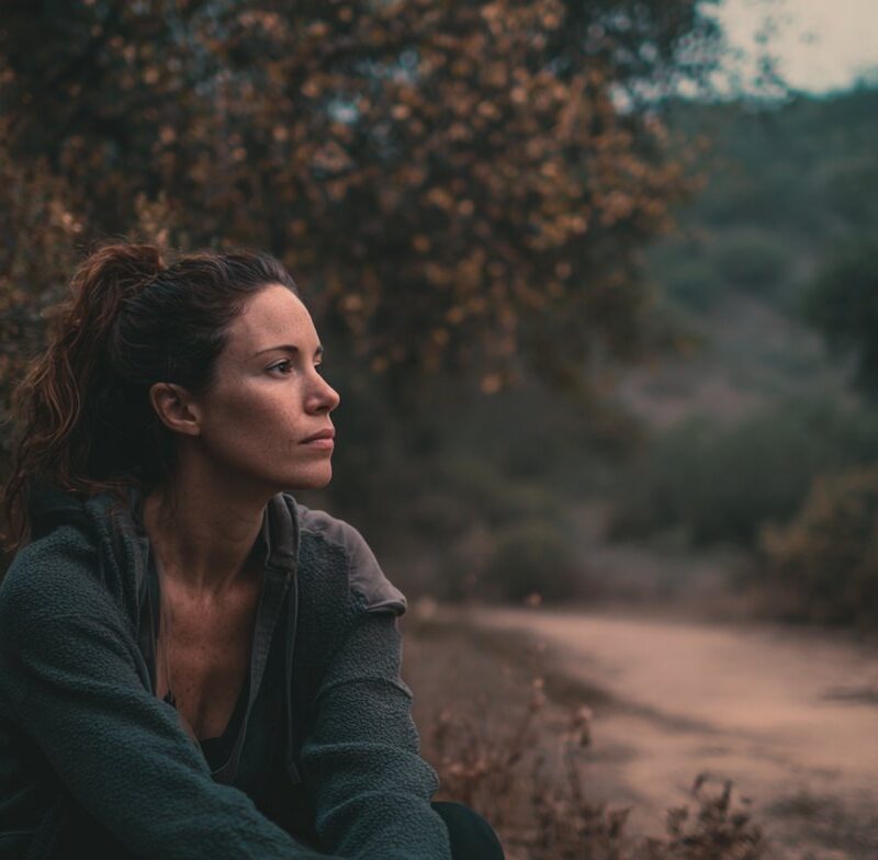 A woman sitting thoughtfully on a forest path at dusk, evoking introspection and healing in trauma therapy that accepts Medi-Cal.