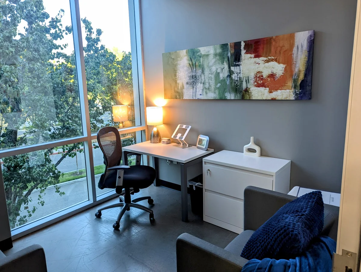 Counseling room at Redwood Counseling’s San Bernardino office, with a desk, upholstered seating, abstract artwork, and windows overlooking trees.